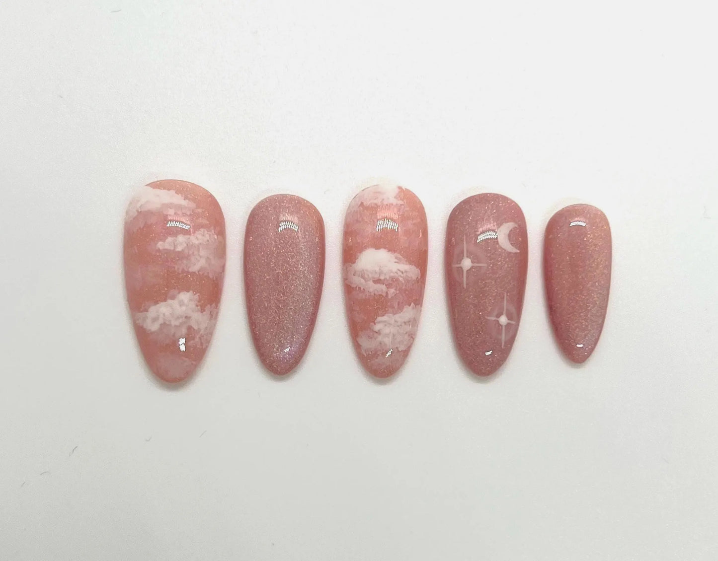 Handmade pink press-on nail samples with cloud and moon designs on a white background