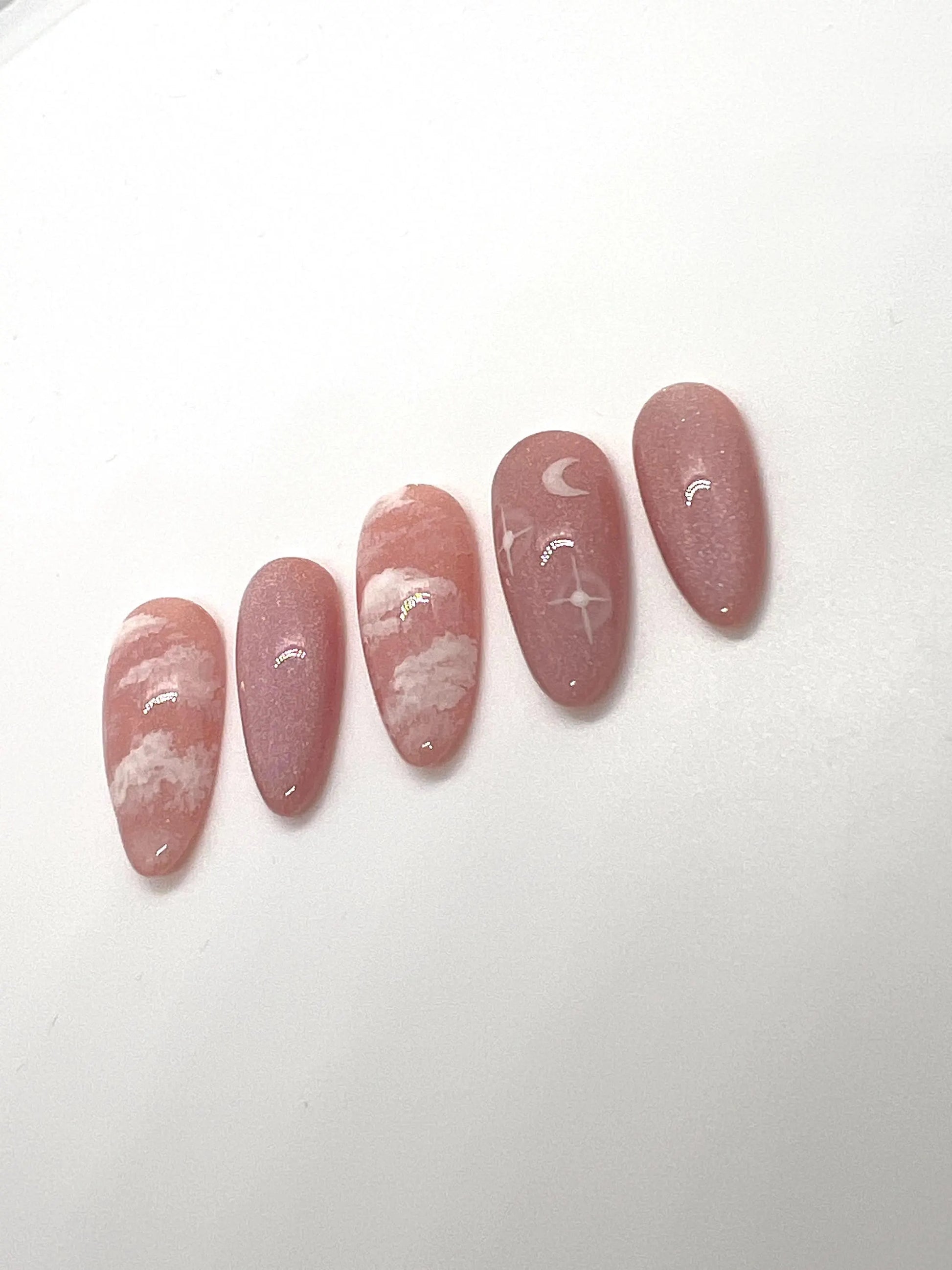 Handmade pink press-on nail samples with cloud and moon designs on a white background